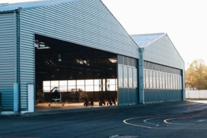 A modern industrial metal hangar exterior with a helicopter visible, offering spacious interior views